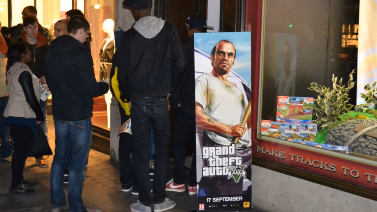 Fila na porta de uma loja de games em Londres na noite de lançamento do GTA V, ao lado do cartaz do jogo com a data 17 de setembro