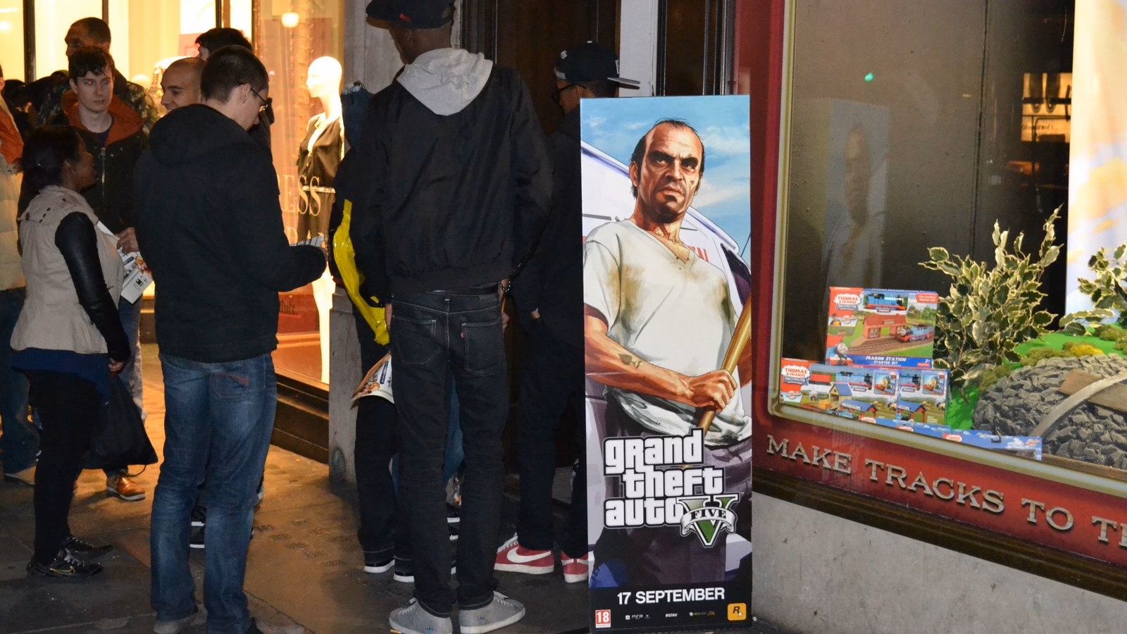 Fila na porta de uma loja de games em Londres na noite de lançamento do GTA V, ao lado do cartaz do jogo com a data 17 de setembro